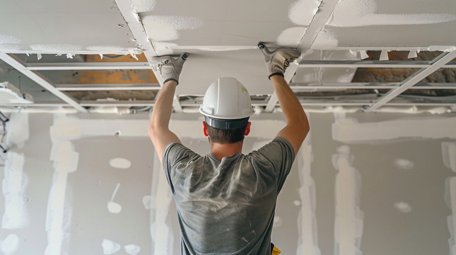 A construction worker in a hard hat and gloves installs drywall on a ceiling in a room with unfinished walls, conveying focus and craftsmanship.