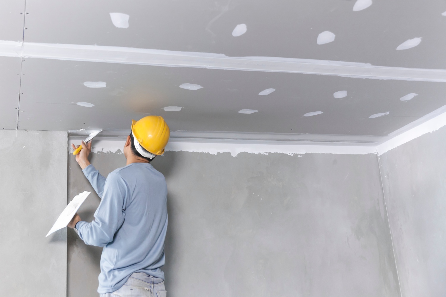 A construction worker wearing a yellow hard hat applies joint compound to drywall seams on a ceiling and wall to smooth and prepare the surface for painting.