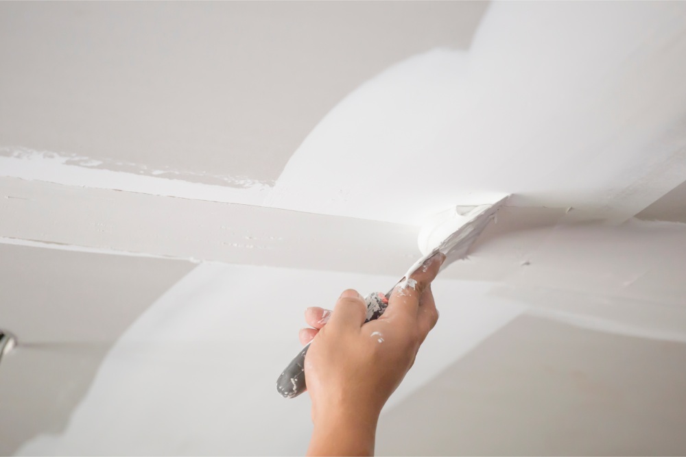 A hand holding a spatula applies white plaster to a ceiling, smoothing the surface at the corner. The setting conveys a sense of focus and precision.