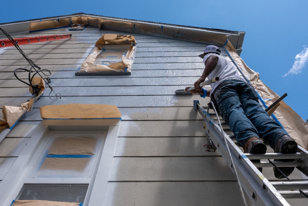Person on ladder painting house exterior light blue under clear sky; windows and roof edges masked with paper, conveying focus and diligence.