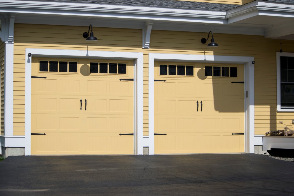Yellow house exterior with two side-by-side garage doors, each having small windows at the top. Black lantern-style lights hang above, casting a welcoming tone.
