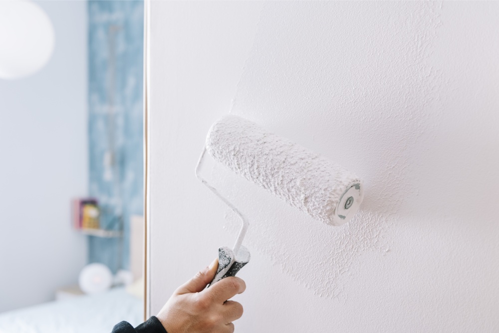 A hand uses a paint roller to apply white paint on a wall, with a blurred background showing a room with soft blue tones. The scene conveys calmness.