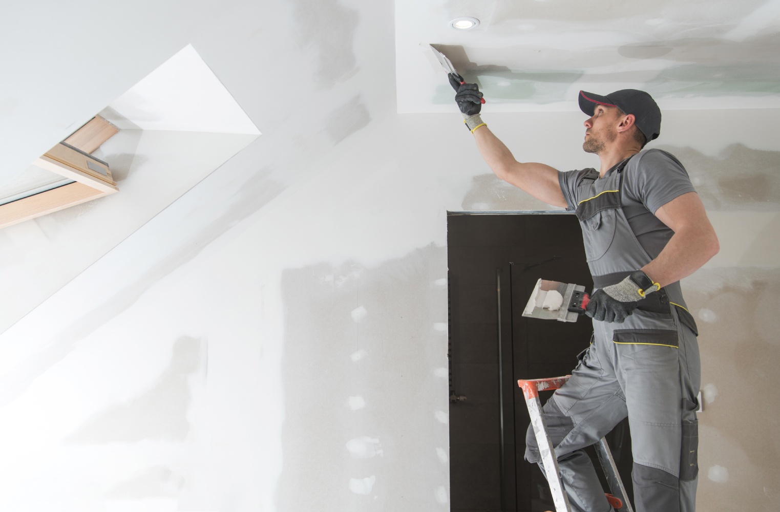 Man on ladder plastering a ceiling in a room with slanted walls. He wears work gloves and gray overalls, concentrating on the task. Bright, clean setting.