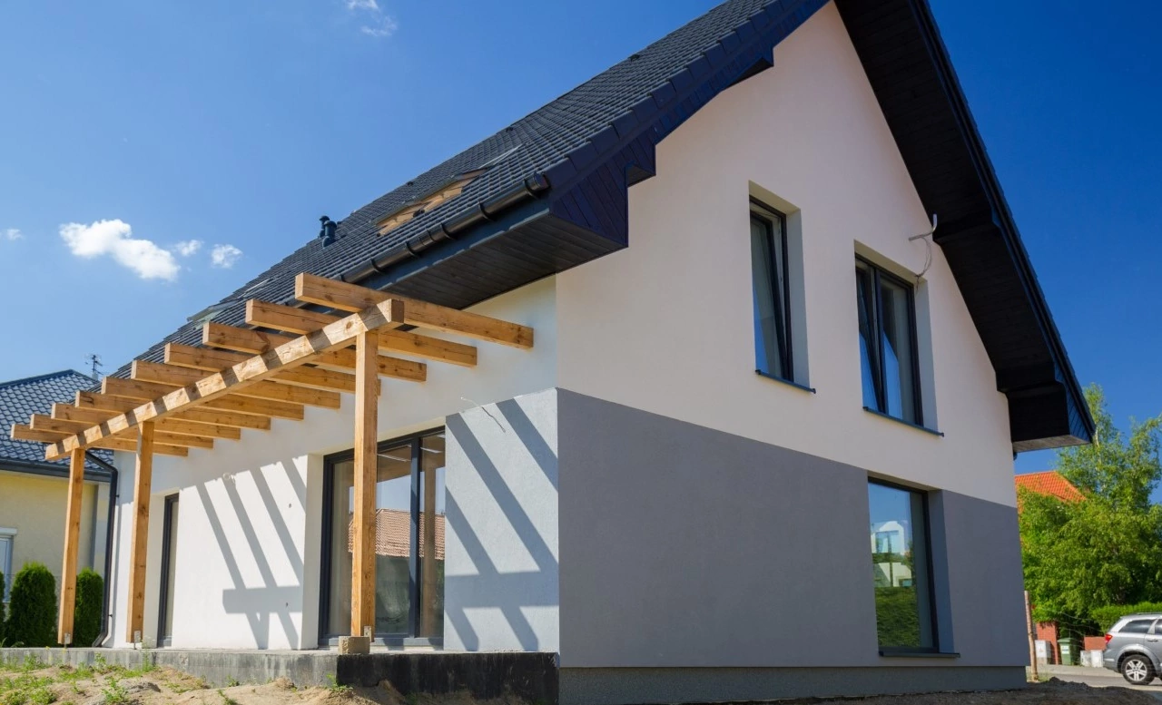 Modern house with a sloped roof and large windows, featuring a wooden pergola casting shadows. Clear blue sky and greenery create a serene atmosphere.