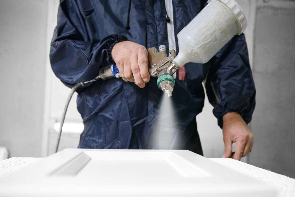 A person in a navy blue protective suit spray paints a white panel in a workshop, showcasing craftsmanship and concentration on the task.