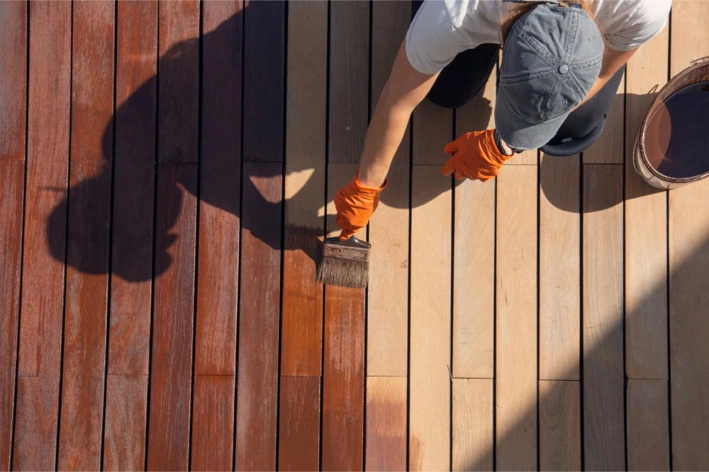 Person in a cap and gloves stains a wooden deck with a brush. The wood transitions from light to dark brown, casting distinct shadows.