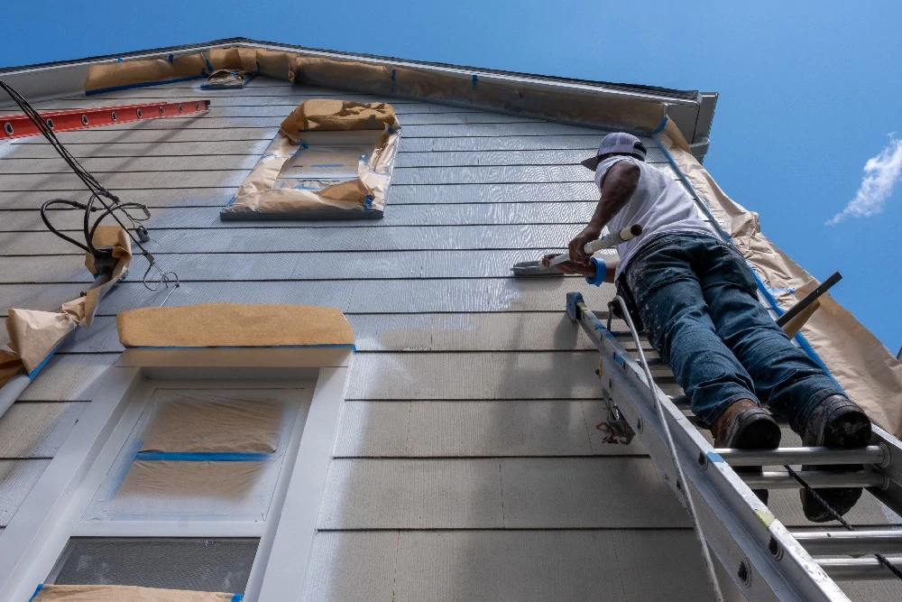 Person on ladder painting house exterior light blue under clear sky; windows and roof edges masked with paper, conveying focus and diligence.