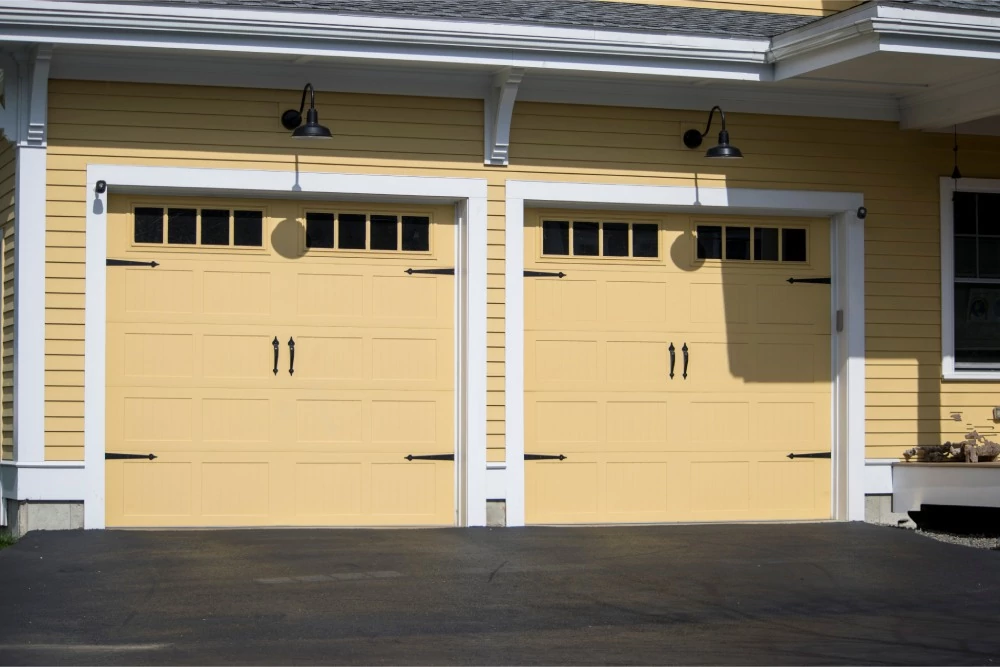 Yellow house exterior with two side-by-side garage doors, each having small windows at the top. Black lantern-style lights hang above, casting a welcoming tone.