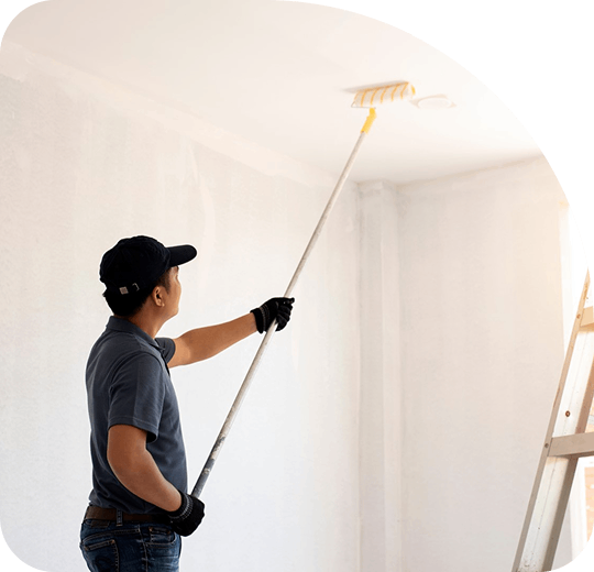 A person wearing a cap and gloves uses a roller with an extended handle to paint a white ceiling. A ladder is visible in the bright room.