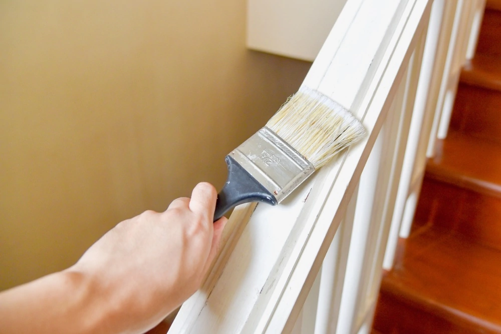A person paints a white staircase railing with a wide paintbrush. The wooden stairs are brown and shiny, creating a neat, home-improvement scene.